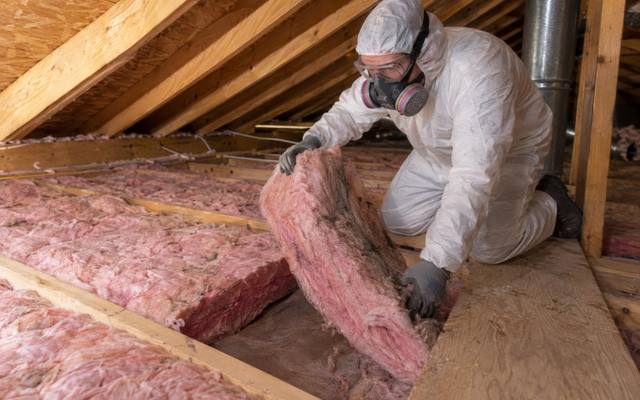 Worker in a white suit and respirator pulling old pink batts out of an attic