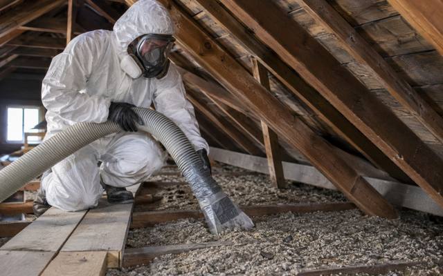 Worker in a protective suit and full respirator holding a vacuum hose over loose pebble-like attic fill