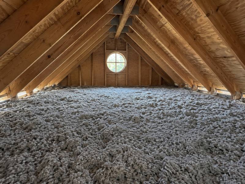 Deep layer of grey blown-in fill across an attic floor below a gable with a round window